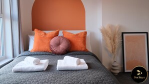 Een net opgemaakt bed met twee opgerolde handdoeken, een groot oranje kussen en een kleiner roze exemplaar. Op het nachtkastje staat een kunstwerk in een lijst en een vaas met pampa gras.