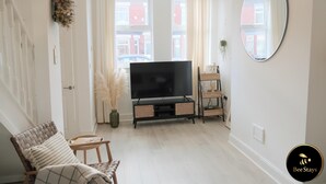 Een woonkamer met een televisie, een stoel, een kleine tafel, een spiegel en een raam met gordijnen.