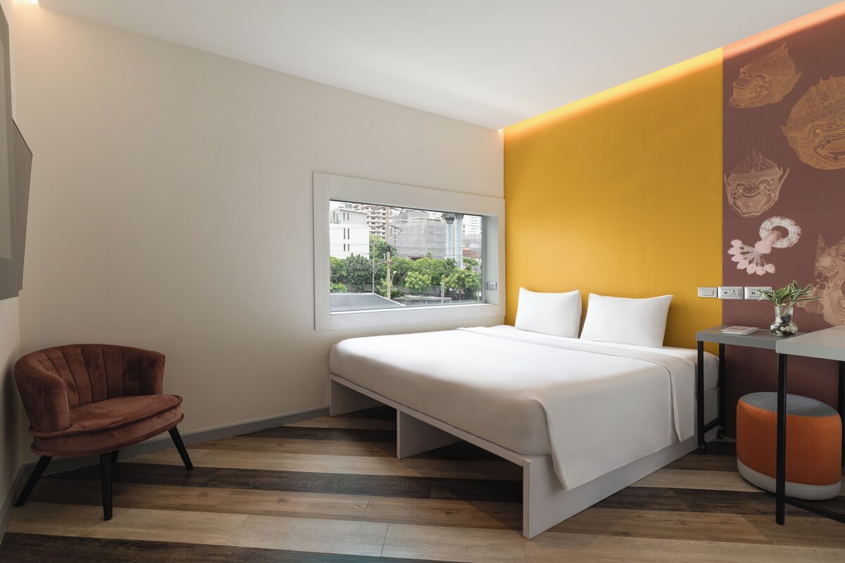 Een moderne hotelkamer met een bed, een stoel, een klein tafeltje met een vaas en een raam met uitzicht op gebouwen.