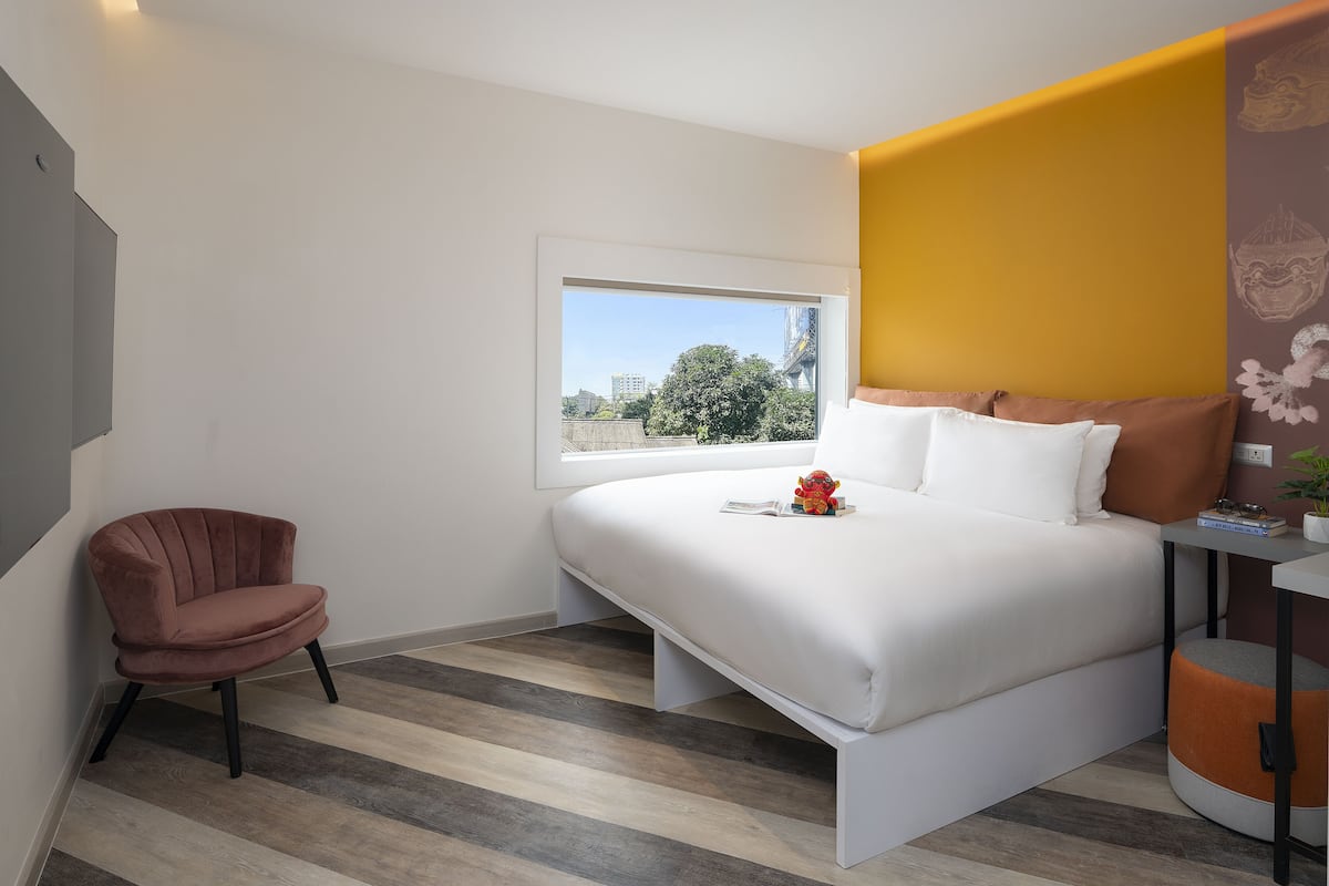 Une chambre d’hôtel moderne avec un grand lit, une fenêtre donnant sur la ville, un fauteuil et une petite table agrémentée d’une plante.