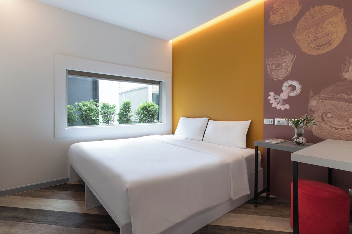 Een moderne hotelkamer met een groot bed, een raam met uitzicht op groen, een nachtkastje met een vaas, en een rode stoel.