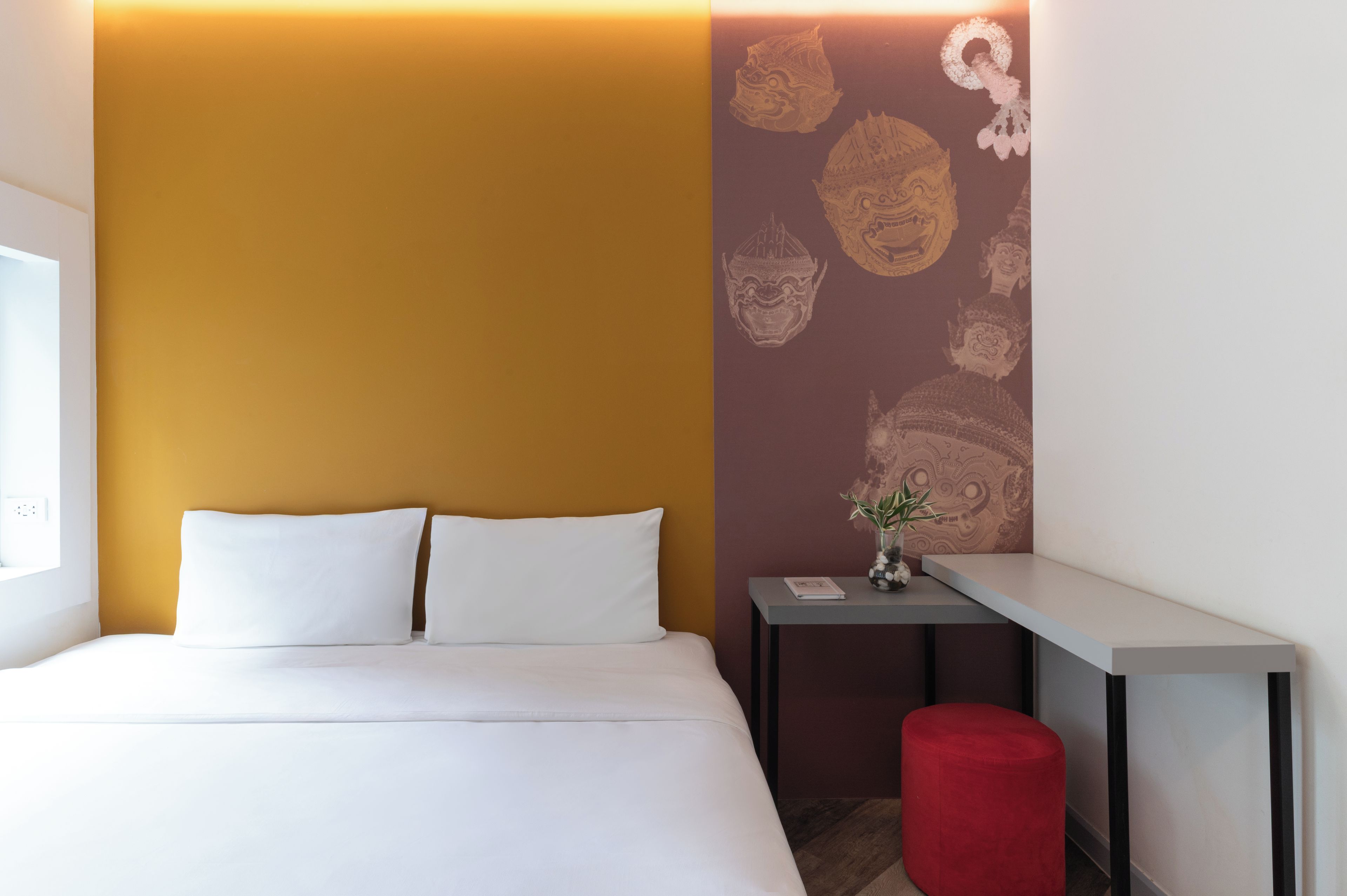 Une chambre d’hôtel avec un lit, une petite table sur laquelle est posé un vase, et un pouf rouge.