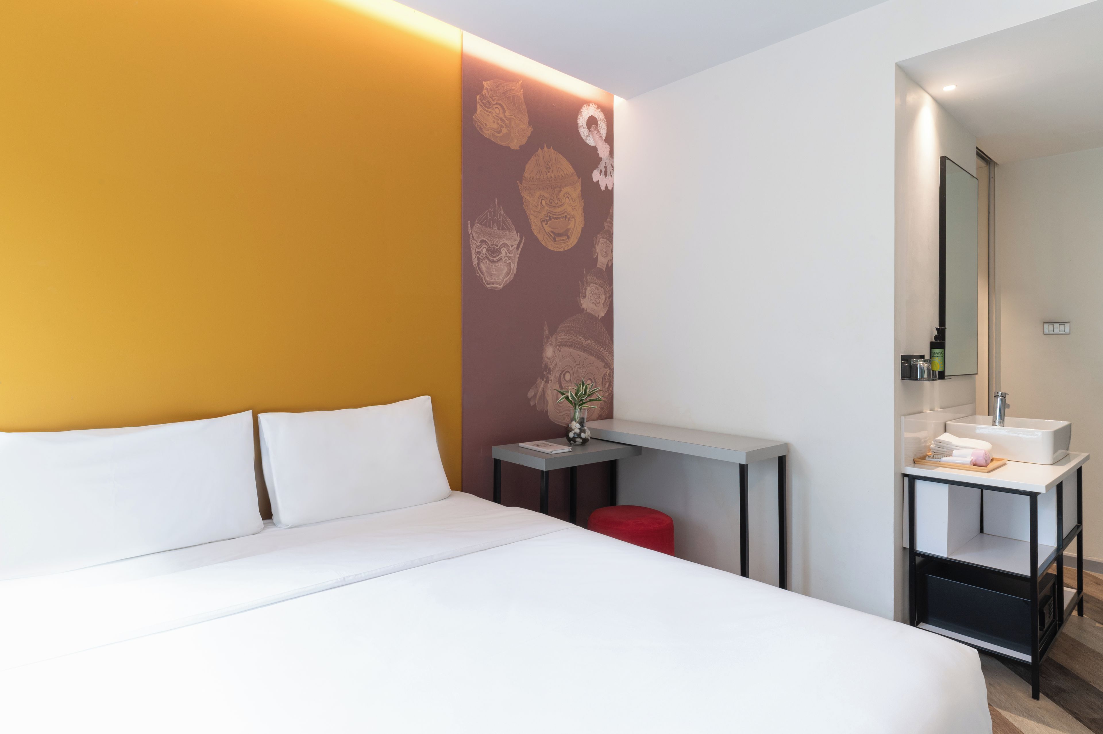 A hotel room with a bed, a small table with a vase, a mirror, and a sink.