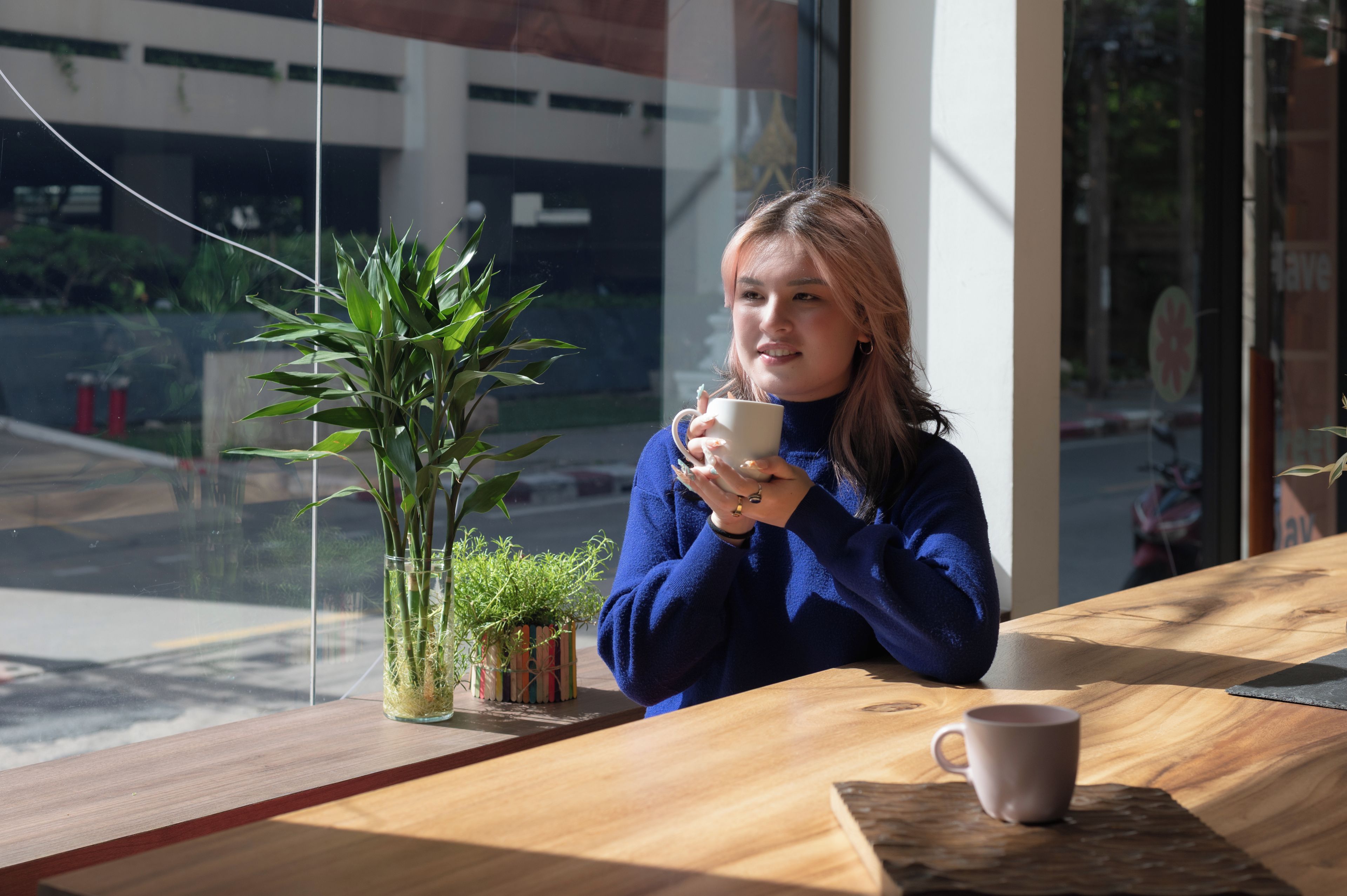 A person sitting at a wooden table with a cup, a potted plant, and a window in the background.