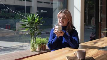 A person sitting at a wooden table with a cup, a potted plant, and a window in the background.