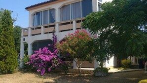 A two-story building with a balcony, surrounded by trees and flowering plants. - Apartments Topić (Privlaka)