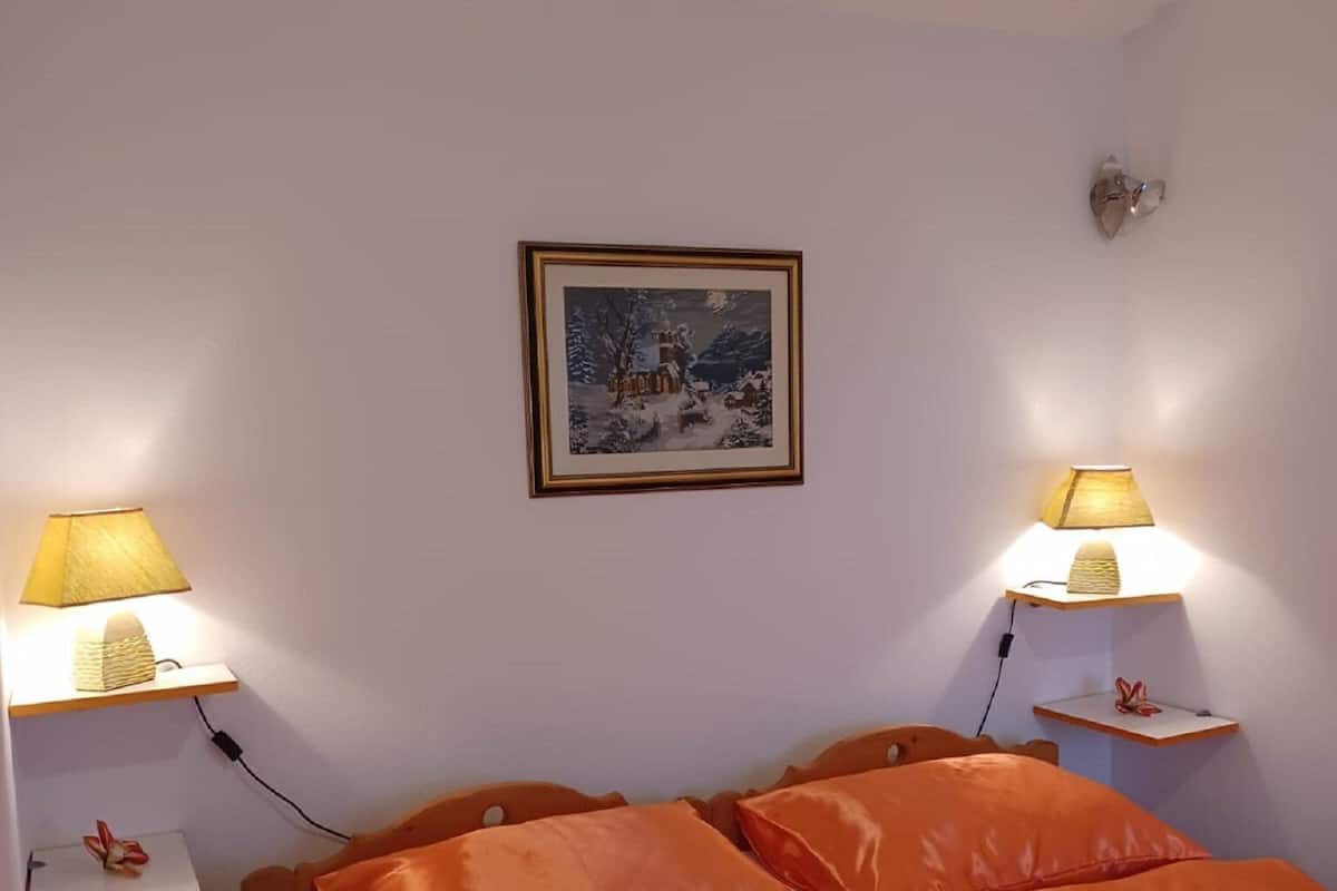 A bedroom with two beds, each with an orange bedspread, two bedside lamps, a framed picture on the wall, and a small shelf with a flower.