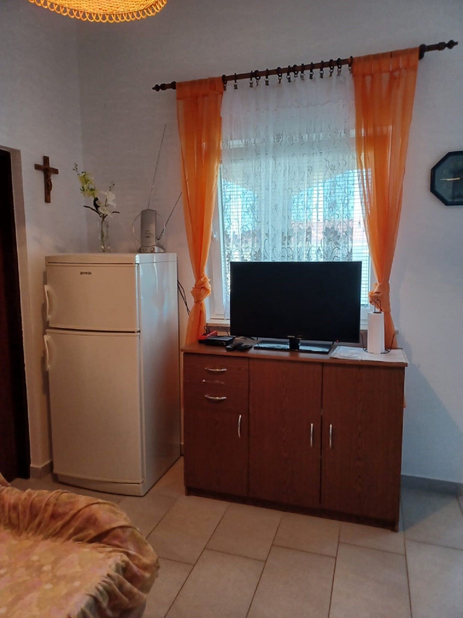 A compact living space with a white refrigerator, a wooden cabinet with a flat-screen TV, and a window with orange curtains.