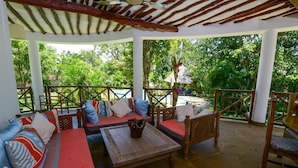 A covered outdoor seating area with a wooden table, cushioned seating, and a view of a pool and lush greenery. - Villa Pundamilia, Private Pool, Free Wifi, Secure (Diani Beach)