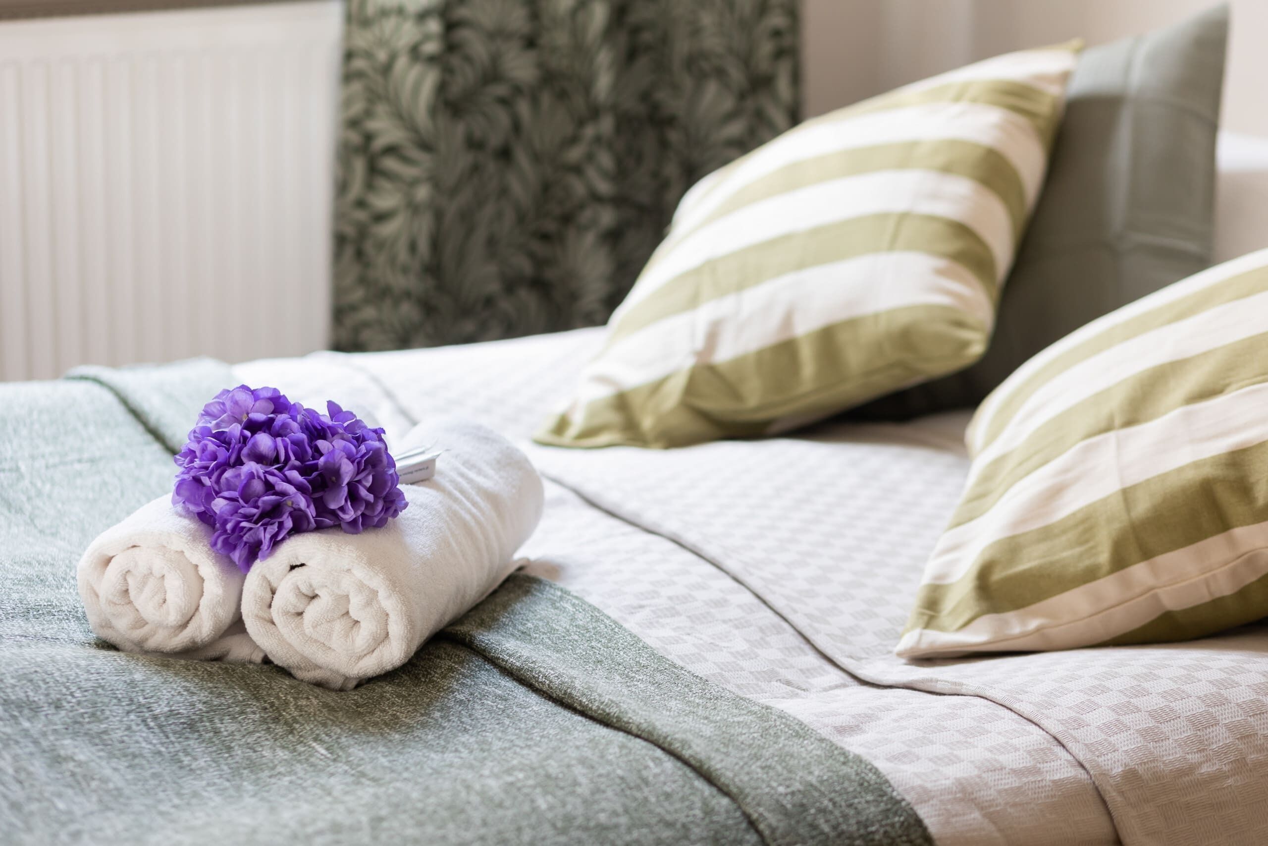 A neatly made bed with two rolled towels and a purple flower, green and white striped pillows, and a patterned green blanket.