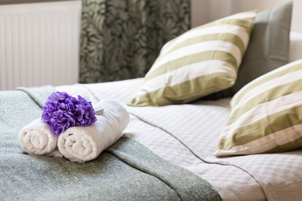 A neatly made bed with two rolled towels and a purple flower, green and white striped pillows, and a patterned green blanket.