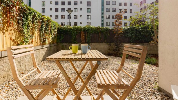 Un espace extérieur aménagé avec une table et des chaises en bois, deux tasses et, en arrière-plan, un bâtiment doté de nombreuses fenêtres et un arbuste.