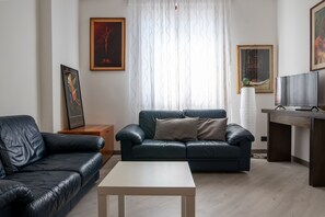 A modern living room with a dark leather sofa, a white coffee table, and a wooden side table. There is a framed artwork on the wall and a television on a wooden stand.