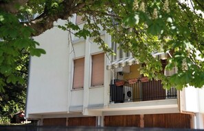 A multi-story building with a balcony, a window with a shutter, and a tree partially obscuring the view.