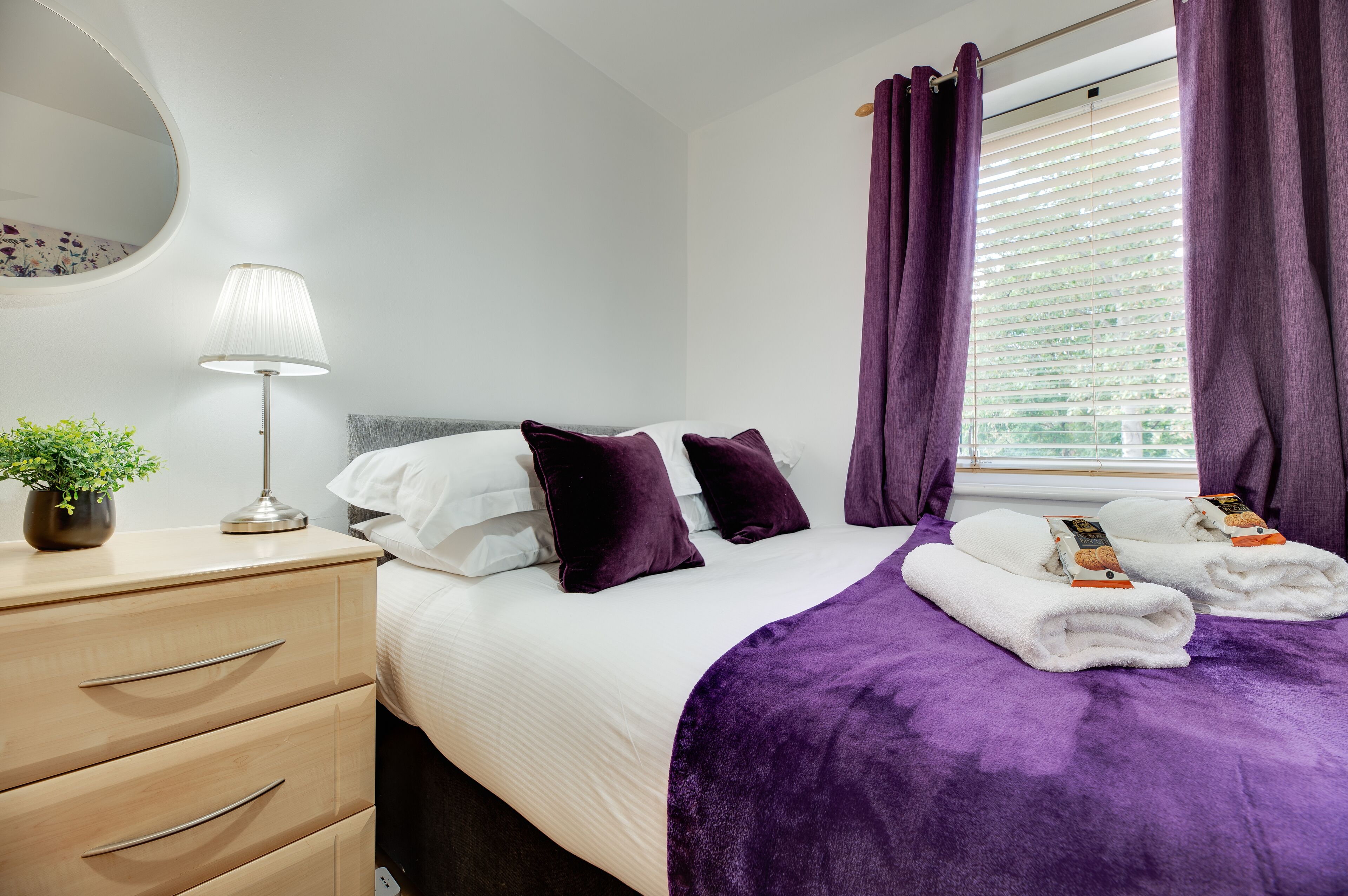 A neatly arranged hotel room with a bed, a bedside table with a lamp, a dresser, and a window with curtains.