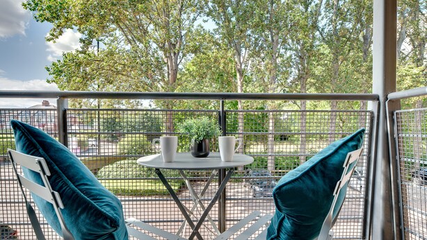 A balcony with two white chairs and teal cushions, a small round table with two white cups and a potted plant, and a view of a green park with trees and a fence. - Week2Week Gosforth apartment w balcony (Newcastle-upon-Tyne)