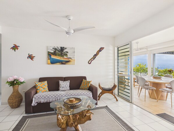 A modern living room with a glass coffee table, a brown sofa, decorative pillows, a ceiling fan, and a painting of a boat.