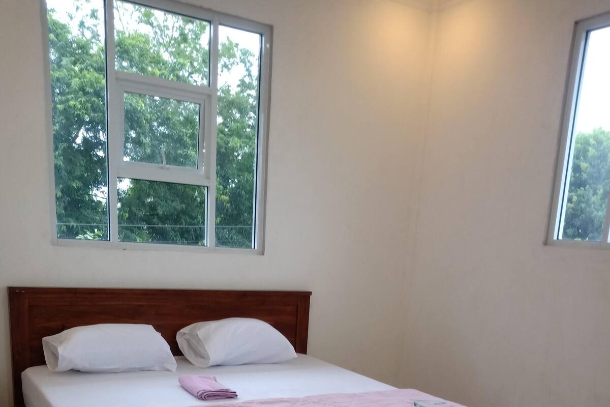 A neatly made bed with a pink floral bedspread, white pillows, and a wooden headboard in a room with large windows and a tiled floor.