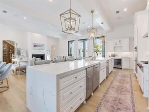 A modern kitchen with a large island, pendant lighting, and a view to the living area.