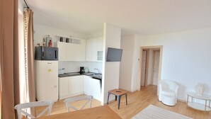 A modern kitchen with white cabinets, a refrigerator, and a microwave. There is a small table and a chair in the dining area. A television is mounted on the wall. - Apartment Borgo Montagna With Pool (San Zeno di Montagna)