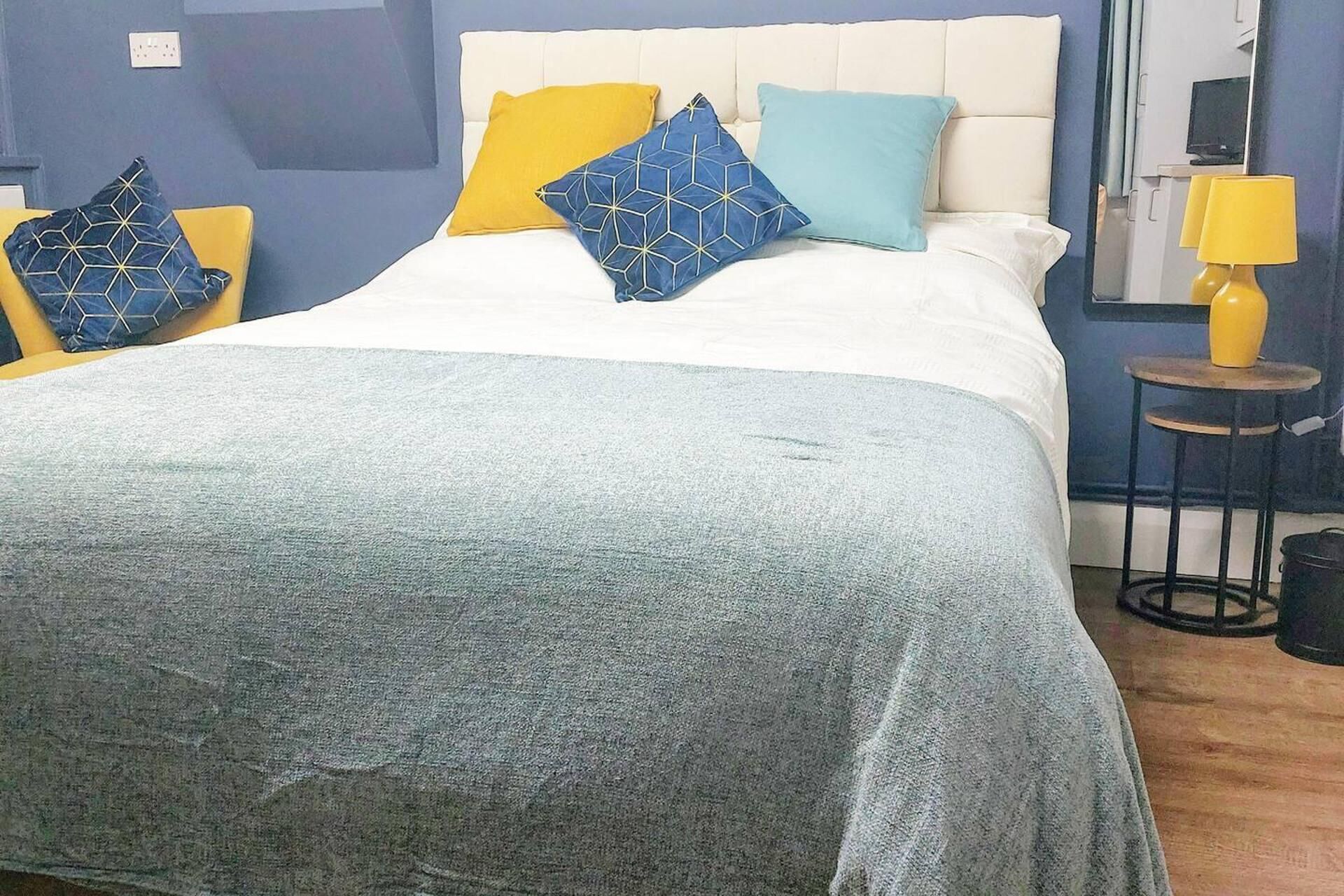 Uma cama bem arrumada com cabeceira branca, manta cinza e várias almofadas em amarelo e azul. Há uma mesa pequena com abajur amarelo ao lado da cama.