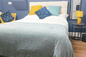 A neatly made bed with a white headboard, a grey throw blanket, and multiple pillows in yellow and blue. There is a small table with a yellow lamp beside the bed.