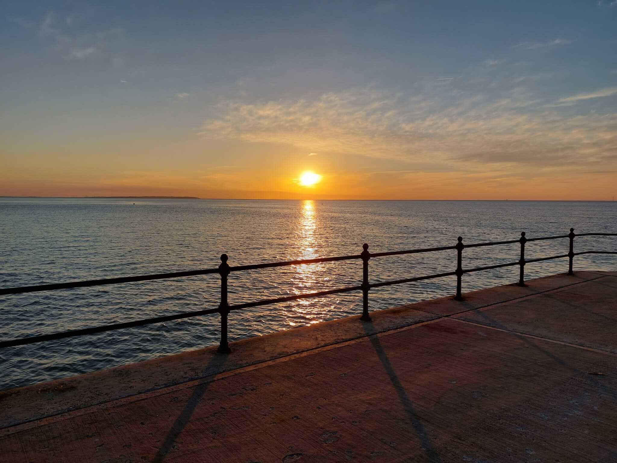 Um pôr do sol sobre o oceano, com um calçadão de concreto e grades de metal.