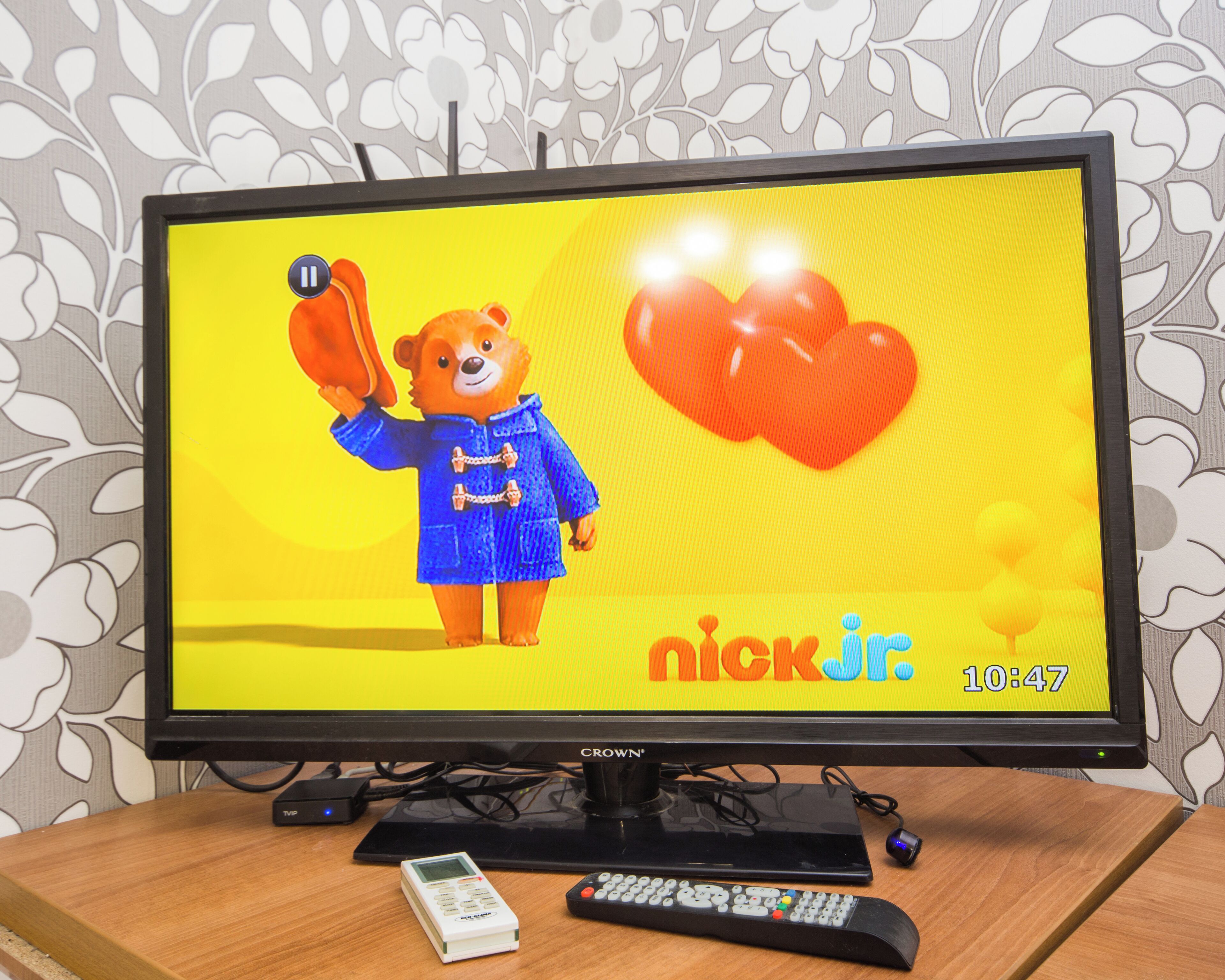 En tv-apparat står på ett träbord och visar en tecknad film från Nick Jr. Bredvid tv:n ligger en fjärrkontroll och en TVP-enhet.