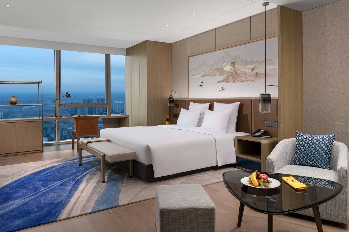 A modern hotel room with a large bed, a seating area with a sofa and armchair, a small table with fruit, and a city view through large windows.
