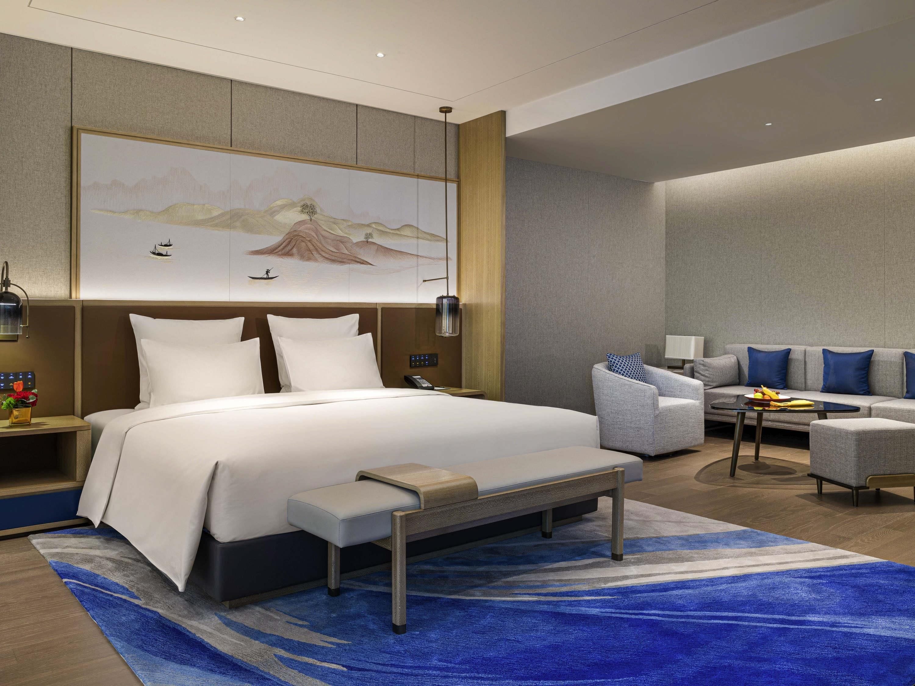 A modern hotel room with a large bed, a seating area with a sofa and armchairs, a small table, and a painting on the wall.