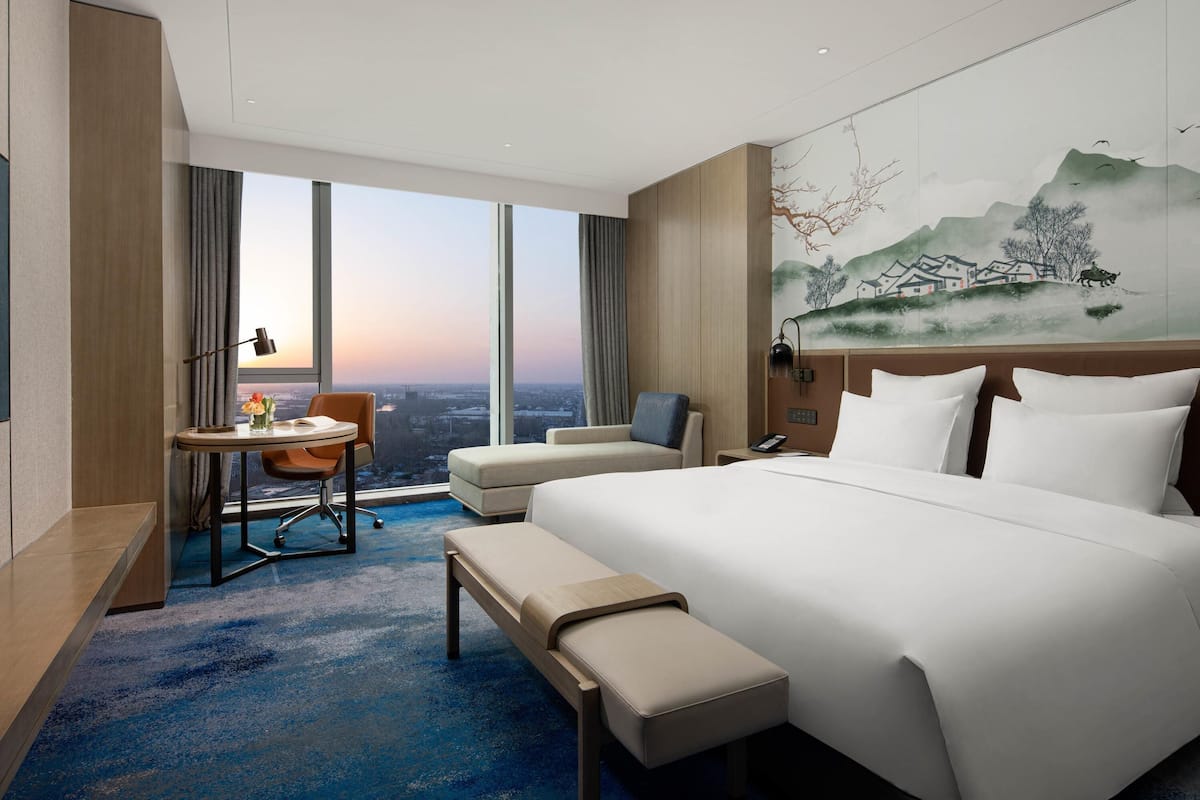 Ein modernes Hotelzimmer mit einem großen Bett, einem Schreibtisch mit Stuhl, einer Bank und Blick auf die Stadt durch große Fenster.