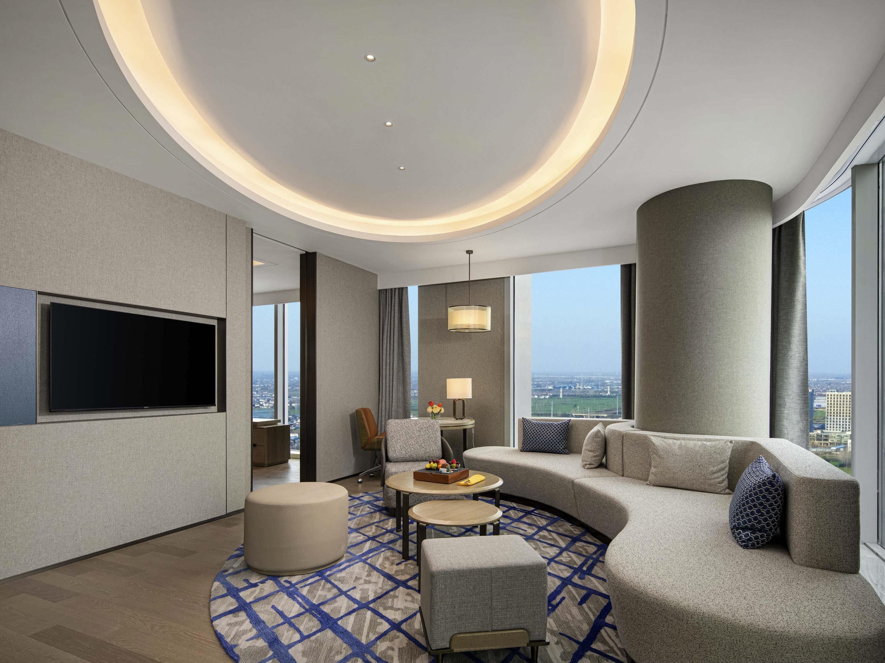 A modern hotel room with a curved sofa, a flat-screen TV, a round coffee table, and a view of the city.