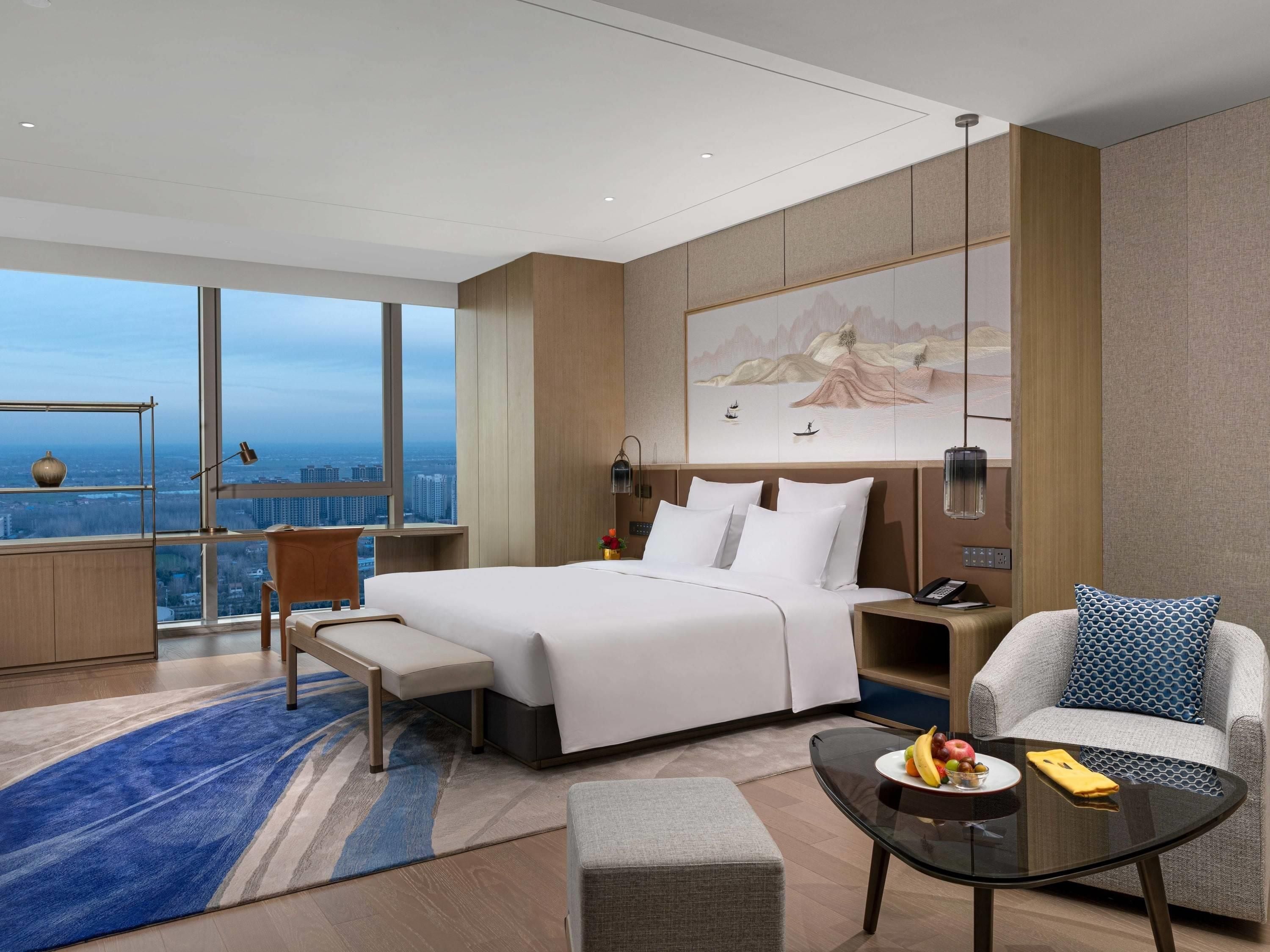 A modern hotel room with a large bed, a seating area with a sofa and armchair, a small table with fruit, and a city view through large windows.