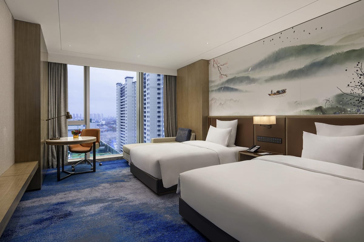 Ein Hotelzimmer mit zwei Betten, einem Schreibtisch und einem großen Fenster mit Blick auf die Stadt.