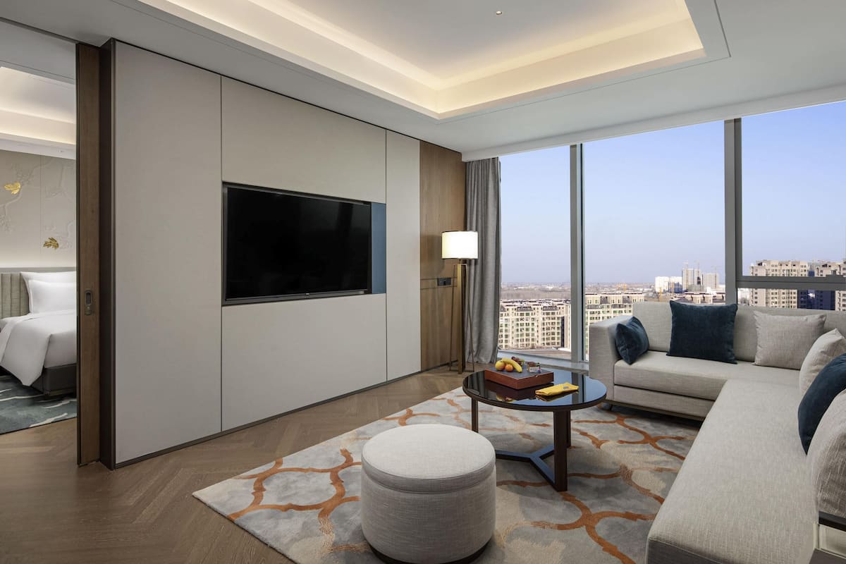 Ein modernes Hotelzimmer mit großem Fernseher, einer Couch, einem runden Couchtisch und Blick auf die Stadt.