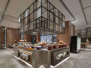 Un hall d’hôtel moderne doté d’un espace buffet central, d’un sol en marbre et d’un plafond haut orné d’une structure géométrique en verre.