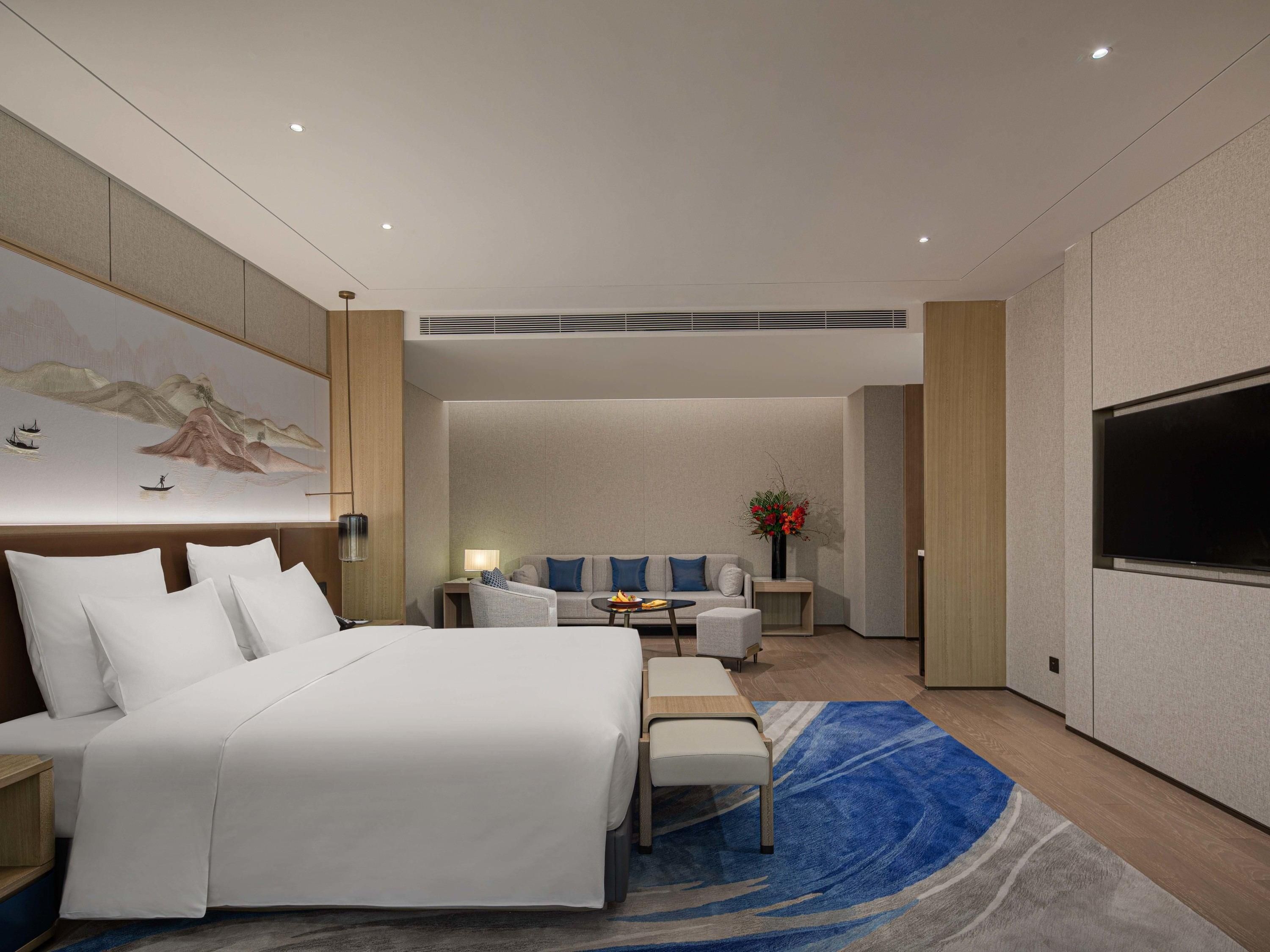 A modern hotel room with a large bed, a seating area with a sofa and chairs, a flat-screen TV, and a decorative wall piece.