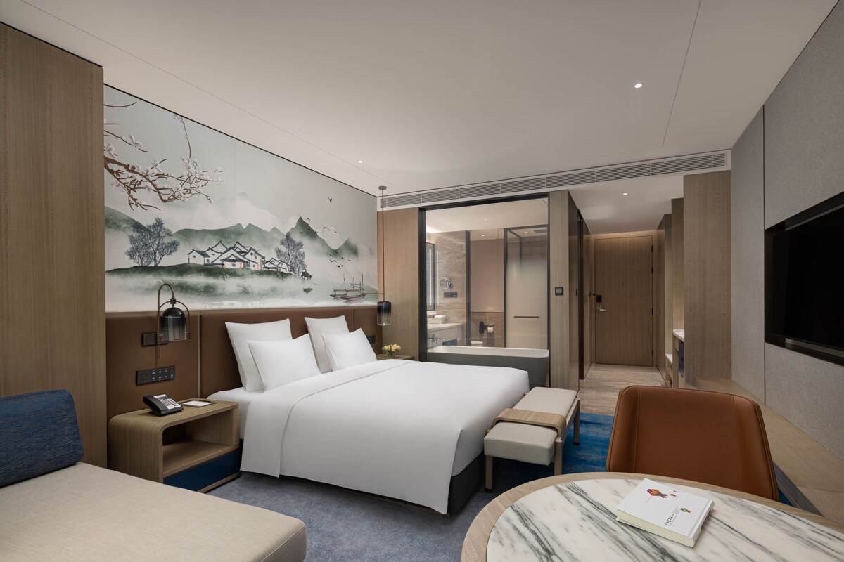 Ein modernes Hotelzimmer mit einem großen Bett, Nachttischen, einem Schreibtisch, einem Sessel, einem kleinen Tisch und einem Gemälde an der Wand.