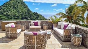 A patio with wicker furniture and stone walls, overlooking a lush green hill and the ocean. - Coulibri Ridge (Soufriere)