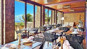 A restaurant with multiple tables set for dining, large windows offering a view of the outdoors, and a stone wall in the background. - Coulibri Ridge (Soufriere)