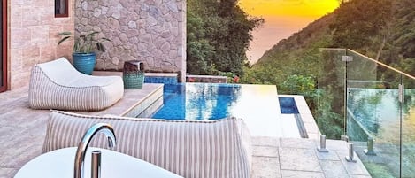 A modern outdoor pool area with a stone wall, a glass railing, and a view of a valley at sunset.