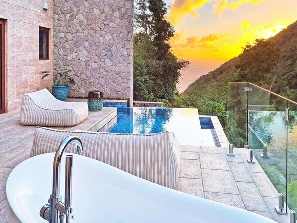 A modern outdoor pool area with a stone wall, a glass railing, and a view of a valley at sunset. - Coulibri Ridge (Soufriere)