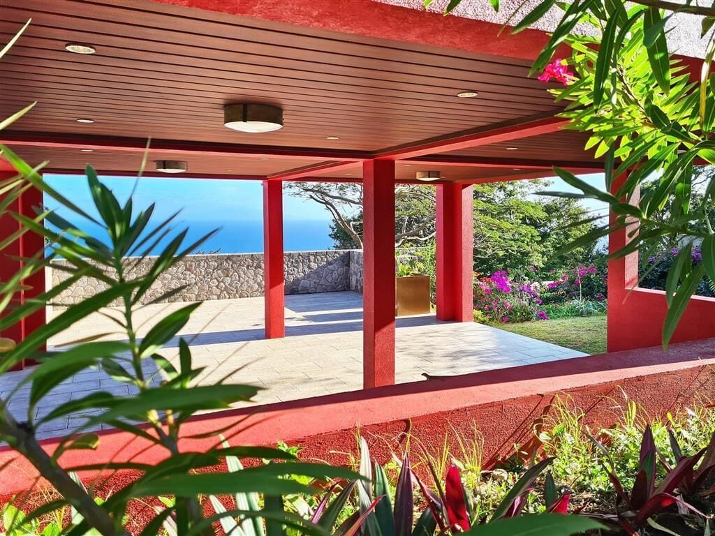 Un espace extérieur abrité, offrant une vue sur l’océan, avec des éléments structurels rouges et diverses plantes.