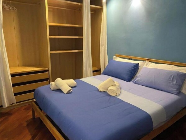 A bedroom with a bed, blue bedding, a wooden wardrobe, and a white curtain.