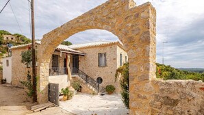 A stone archway leading to a traditional stone house with a tiled roof, a small balcony, and a staircase. - Serenity Villa Retreat in Pidasos (Pylos-Nestoras)