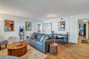 A modern living room with a blue sofa, wooden coffee table, and a dining area with a table and chairs. There are posters on the walls and a ceiling light fixture.