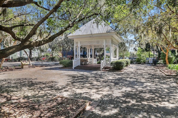 A gazebo with a lattice roof and steps leading up to it, surrounded by trees and a gravel path.