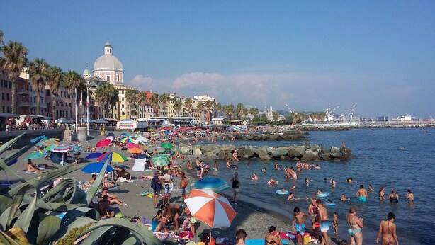 Une plage bondée où l’on voit des gens bronzer et se baigner, des parasols colorés et, au loin, un grand bâtiment surmonté d’un dôme.