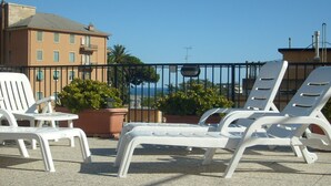 Une terrasse avec des fauteuils de jardin blancs et un pot de fleurs, d’où l’on aperçoit un immeuble et un ciel dégagé.