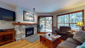 A cozy living room with a stone fireplace, a television mounted on the wall, a comfortable sofa, and a large window with a view of a snowy landscape. - Aspen #120 Prime Ski-in Ski-out Location! Pool, Hot tubs, BBQ, sleeps 6 (Whistler)